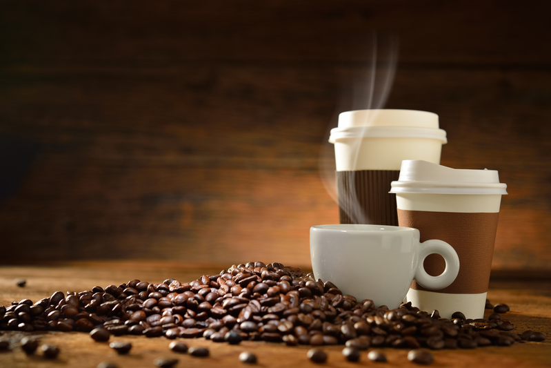 pile of coffee beans in front of two sizes of to-go coffee cups and a white mug